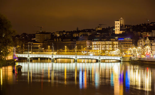 Illuminated bridge over river by buildings against sky at night