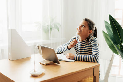 Happy pensive schoolgirl sits at the table at home, does homework on the tablet 