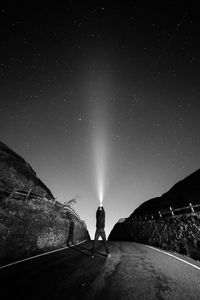 Rear view of man holding illuminated flashlight while standing on road against sky at night