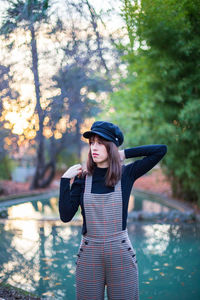 Thoughtful young woman standing against lake at park during sunset