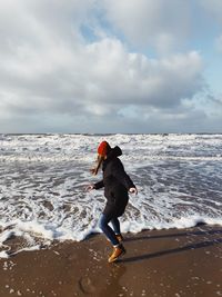 Full length side view of woman wearing warm clothing while playing with waves on shore at beach against sky