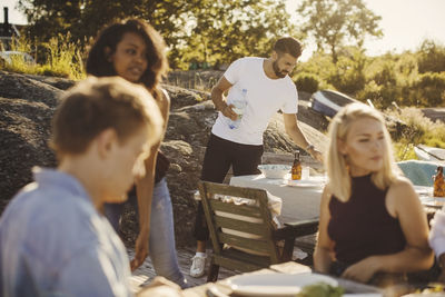 Young multi-ethnic friends sitting while man setting table at harbor on sunny day