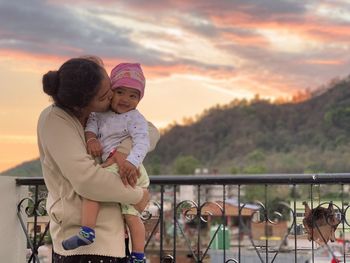 Mother kissing baby girl while standing against sky at sunset