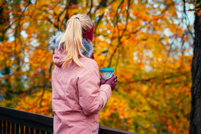 Rear view of woman holding coffee cup standing by railing against autumn leaves