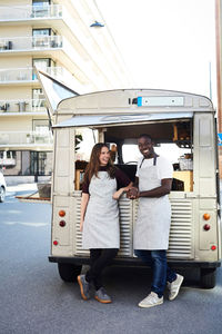 Full length of smiling male and female owners standing outside food truck parked on city street