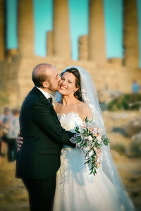 Bridegroom kissing happy bride on cheek during sunset