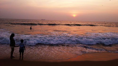 Mother with son standing at seashore against sky during sunset