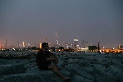 Man sitting on rock by cityscape against sky at night