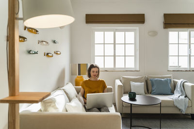 Freelancer working on laptop sitting on sofa in living room at home