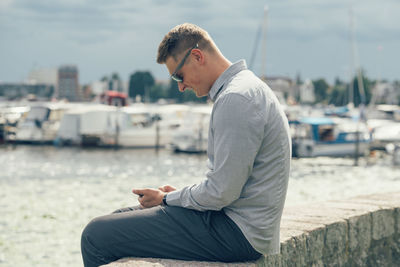 Side view of young man  sitting at the harbor and looking at a mobilephone 