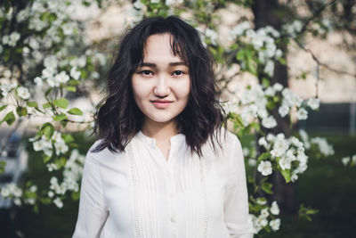 Young attractive asian woman in a white shirt under the blooming tree in park, calm, smiling. spring