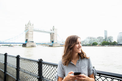 Young woman using phone on bridge against sky