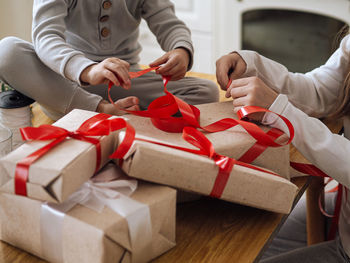 Toddler boy and teenage girl in pajamas packing christmas boxes with red ribbons on the table