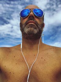 Shirtless mature man wearing sunglasses while listening music against sky