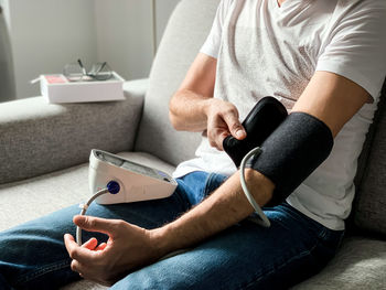 Man checking blood pressure at home. sitting on gray sofa. seasonal issue. arranging monitor on hand