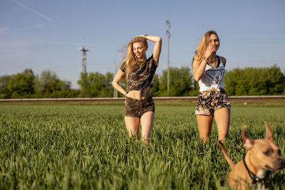 Female friends with dog walking on grassy field against sky