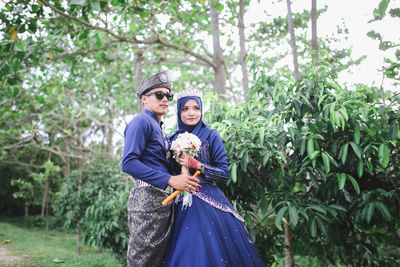 Bride and groom holding flower bouquet while standing by tree