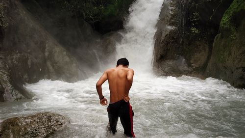 Rear view of shirtless man standing against waterfall