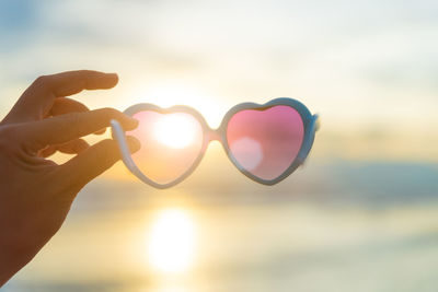 Cropped hand holding heart shape sunglasses against sky
