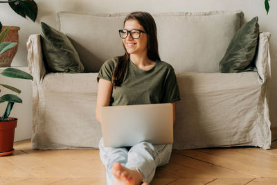 Young happy female in glasses works on a laptop while sitting on the floor in the apartment.