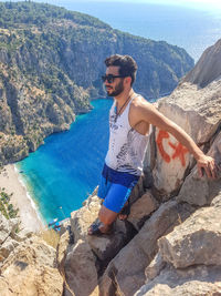 Young man wearing sunglasses while standing on rock over sea against mountains