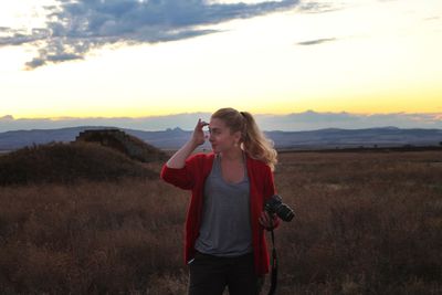 Young woman with camera while standing on landscape against sky during sunset