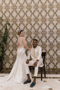 Beautiful bride standing by happy groom smiling while sitting on chair against wall