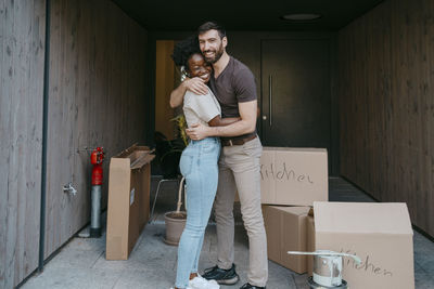 Portrait of happy man embracing girlfriend while standing amidst cardboard boxes