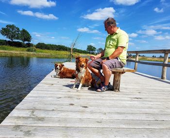 Man with dog sitting by lake against sky