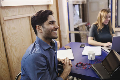 Happy young man looking away while sitting at desk with colleague in board room