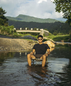 Portrait of young man sitting by lake against sky