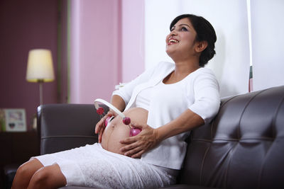 Pregnant woman holding headphones on belly while sitting on sofa at home