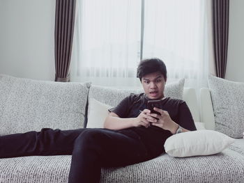 Shocked young man using smart phone while relaxing on sofa at home