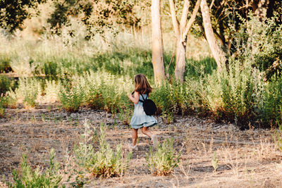 Rear view of girl with backpack running on field against plants in forest