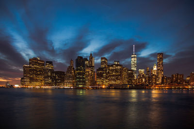 Illuminated one world trade center amidst buildings in front of sea at dusk