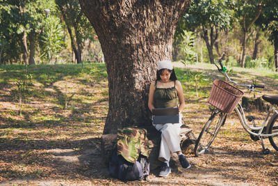 Asian woman in casual wear sitting and working with a laptop under the tree next to a white bicycle