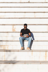 Young man using digital tablet while sitting on staircase during sunny day