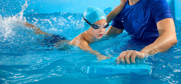 Male trainer teaching preschool boy how to swim in indoor pool with pool flutter board