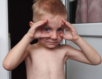 Close-up portrait of cute shirtless boy holding eyes wide open at home