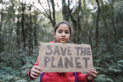 Ethnic child showing save the planet title on carton piece while looking at camera in forest