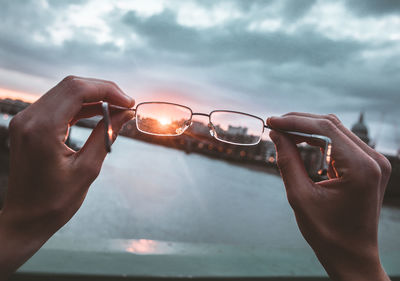 Cropped hands of person holding eyeglasses against sky