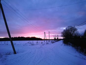 Electricity pylons on snow covered field against sky during sunset