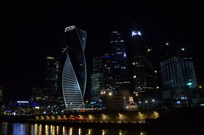 Illuminated modern buildings by river against sky at night