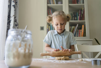 Blond girl rolling cookie dough on table at home