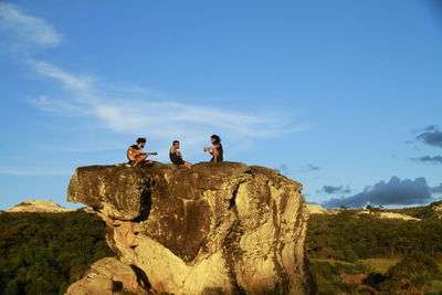 Friends sitting on rock formation against sky