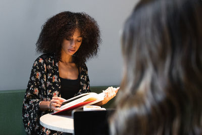 Young ethnic female with afro hairstyle reading textbook at cafeteria table against crop unrecognizable friend