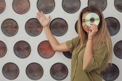 Portrait of woman looking through compact disc while standing against wall