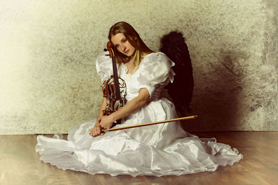 Young woman in angel costume sitting on floor with violin against wall