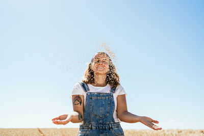 Low angle of delighted female tossing up wheat grain while standing in agricultural field in sunny day in village