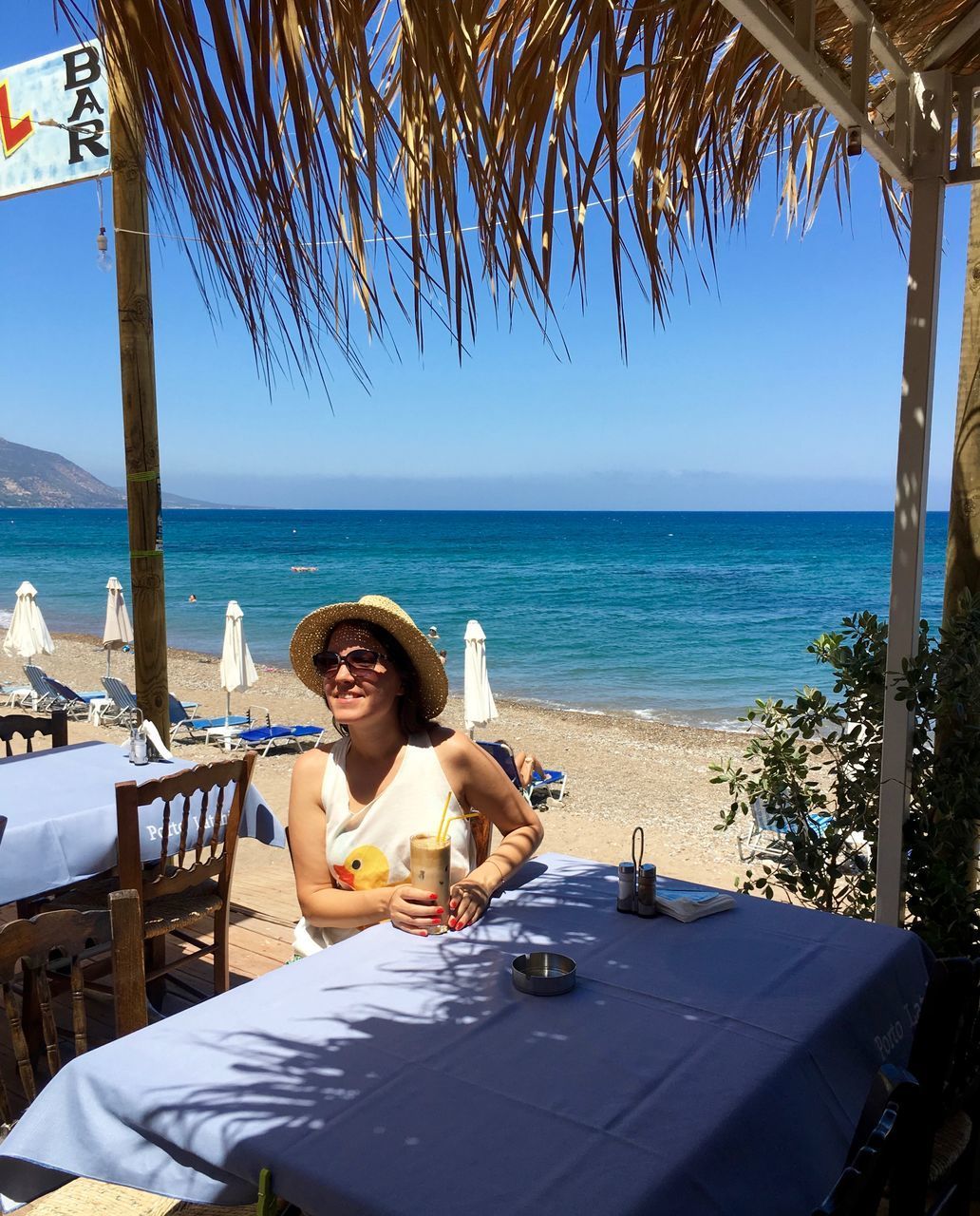 YOUNG WOMAN SITTING ON TABLE BY SEA AGAINST CLEAR SKY